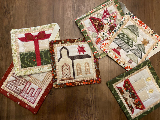 Five handmade quilted fabric coasters with holiday and rustic designs, arranged on a dark wood tabletop.